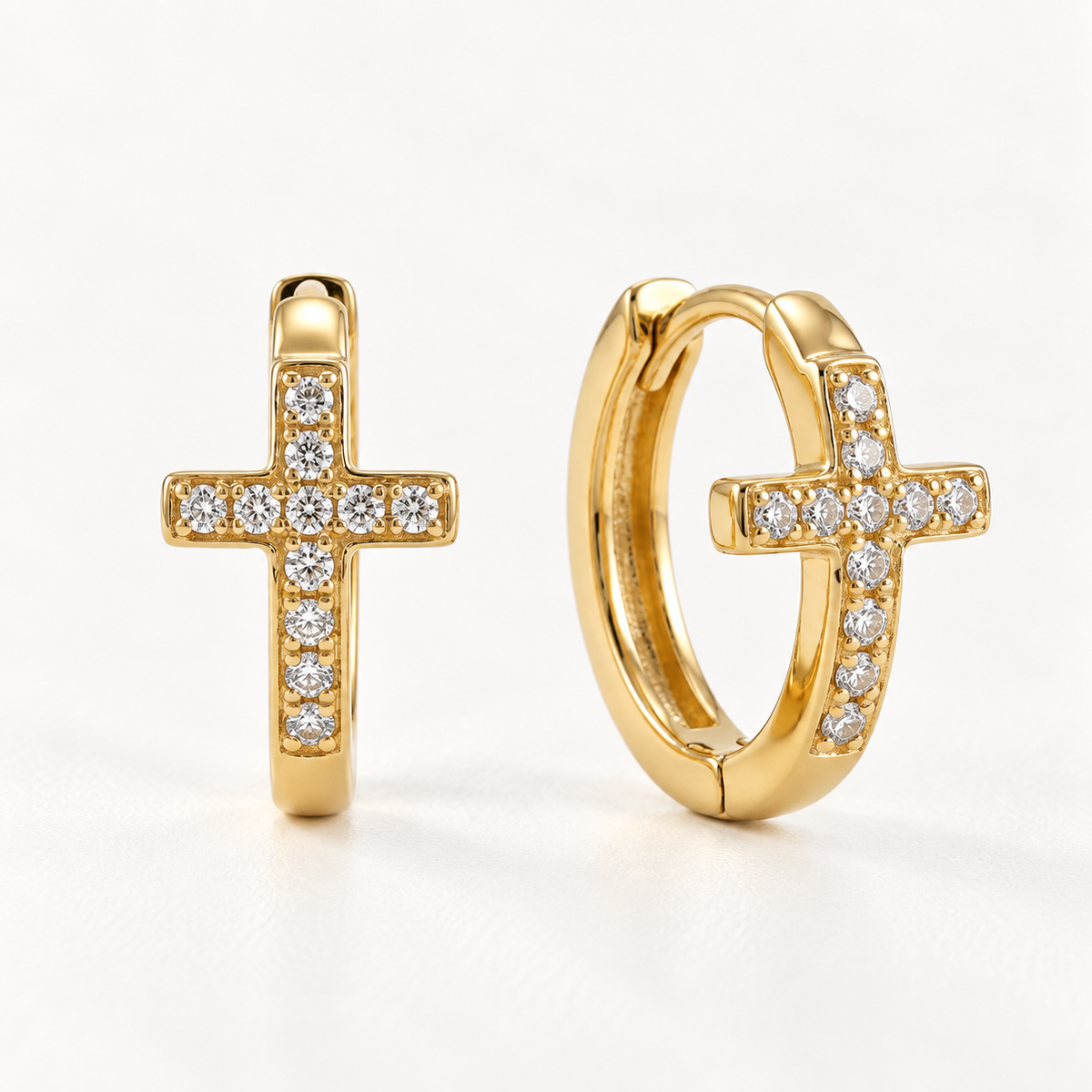 A pair of 18k gold cross-shaped huggie hoop earrings with clear pave crystals along the edges, shown from front and side angles on a clean white background displaying the hinged closure and sparkling stone detail.