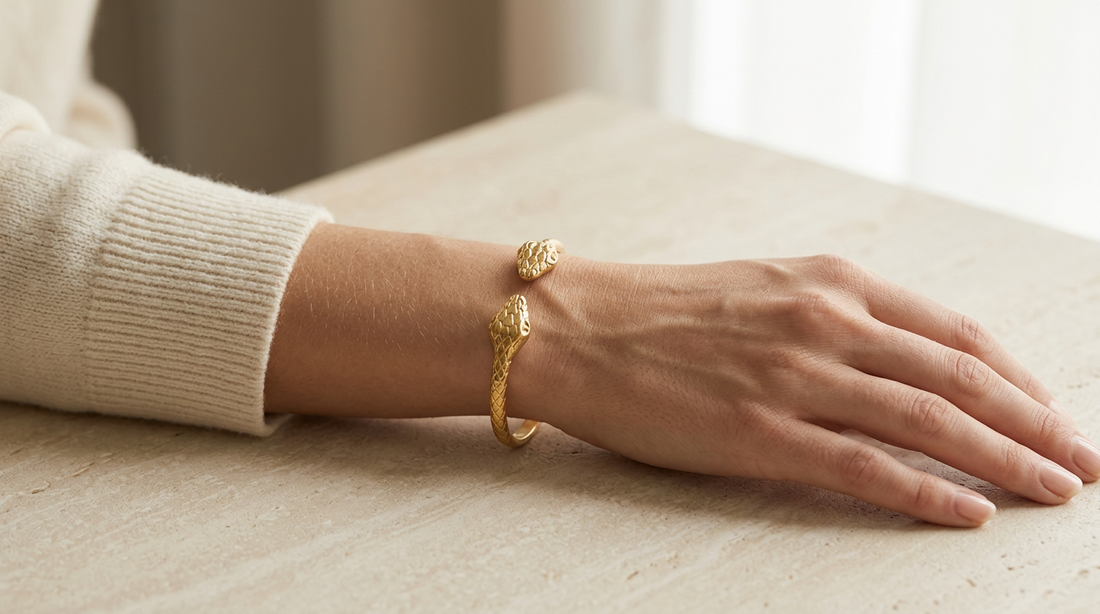 Close-up of a feminine wrist wearing a gold double-headed snake bangle cuff with textured scale pattern, the serpent heads facing outward on either side of the wrist. Soft natural lighting, warm beige linen background, natural skin texture with visible pores and slight imperfections.