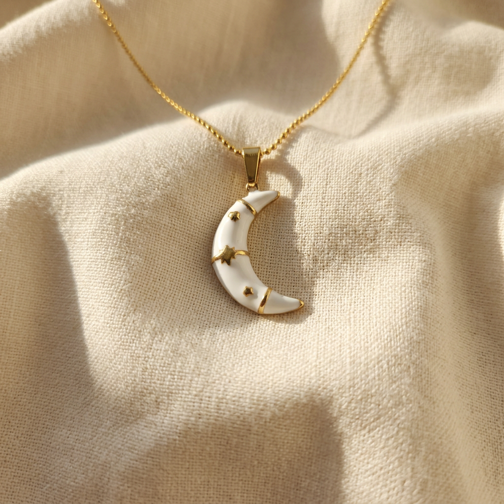A gold enamel moon necklace with white crescent moon pendant and gold star accents lying flat on a white background with soft natural shadows, showing the glossy enamel finish, celestial star detailing, cable chain, and polished gold finish from above.
