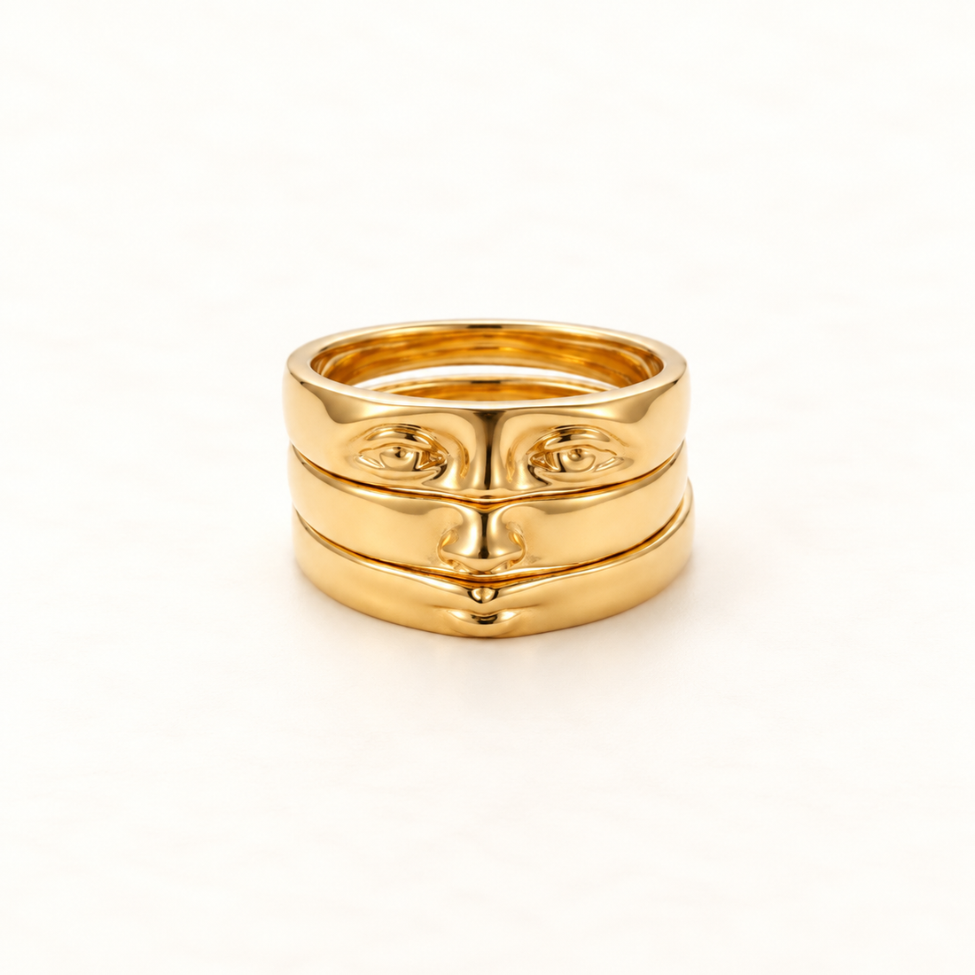 A gold face ring with surrealist facial features including eyes, nose, and mouth across stacked bands, open adjustable design, shown from front and side angles on a clean white background displaying the artistic anthropomorphic detail, sculptural expression, and polished gold finish.