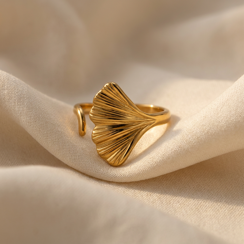 A gold ginkgo leaf ring with detailed vein texture and fan-shaped organic silhouette lying flat on a white background with soft natural shadows, showing the intricate leaf ridges, natural botanical form, and open adjustable band design from above.