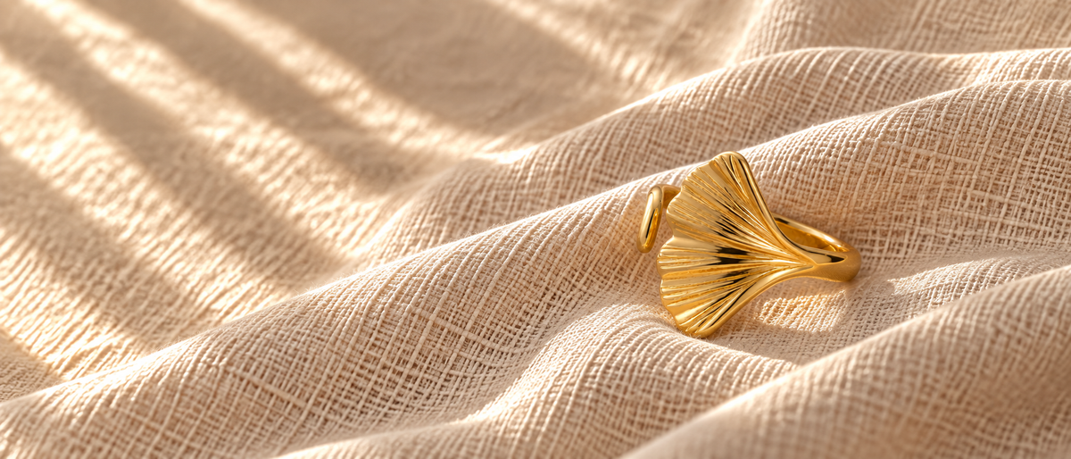 Gold ginkgo leaf ring on oatmeal linen fabric with dappled sunlight creating soft shadows. Warm golden tones, organic texture, intimate jewelry photography.