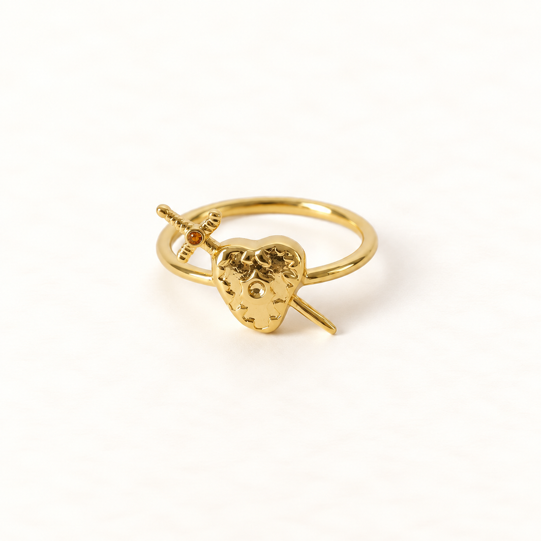 A gold heart and sword ring with detailed anatomical heart pierced by a dagger, open adjustable band, shown from front and side angles on a clean white background displaying the romantic yet edgy symbolism, intricate heart texture and blade detail, and polished gold finish.