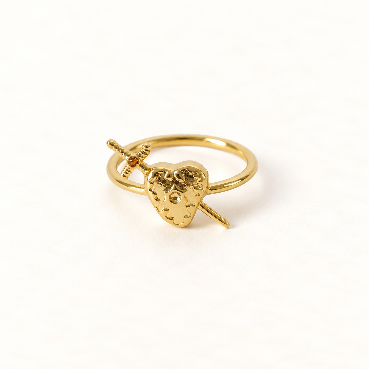 A gold heart and sword ring with detailed anatomical heart pierced by a dagger, open adjustable band, shown from front and side angles on a clean white background displaying the romantic yet edgy symbolism, intricate heart texture and blade detail, and polished gold finish.