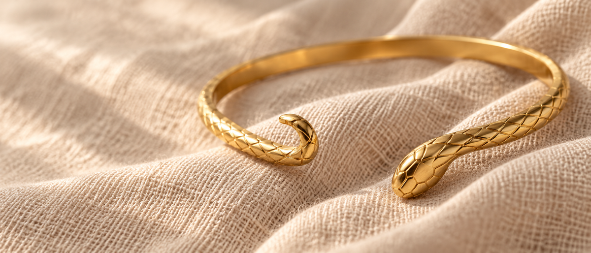 Gold snake bangle bracelet on oatmeal linen with soft dappled lighting. Textured scale pattern and serpent head detail. Warm golden tones.