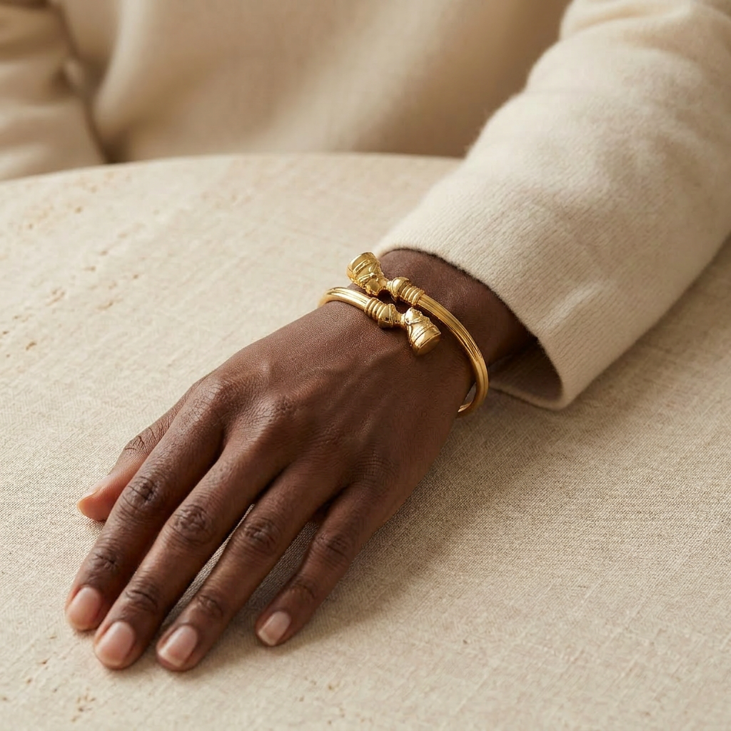 Close-up of a feminine wrist wearing a gold Nefertiti collar bracelet with Egyptian-inspired ribbed texture and ornate cylindrical end caps, the regal cuff resting elegantly on the wrist bone. Soft natural lighting, warm beige linen background, natural skin texture with visible pores and slight imperfections.