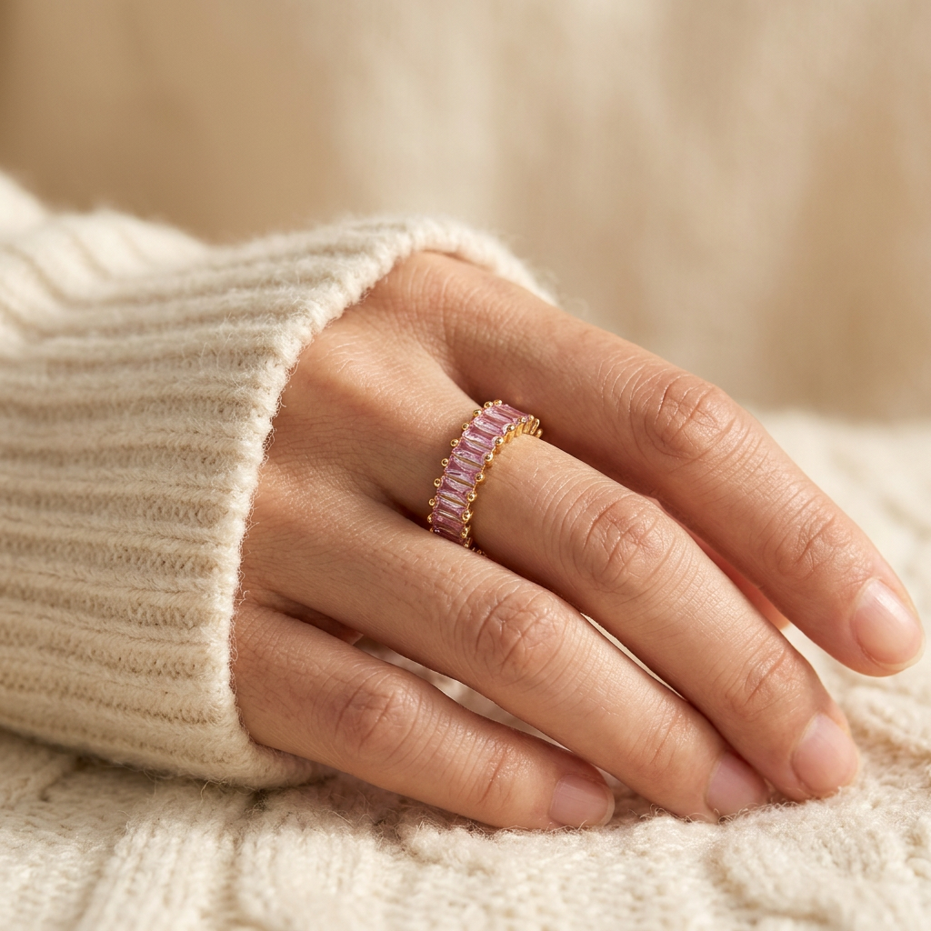 Close-up of a feminine hand wearing a gold pink baguette ring with continuous band of rectangular pink crystals, the soft rose hue sparkling on the finger. Soft natural lighting, warm beige linen background, natural skin texture with visible pores and slight imperfections.