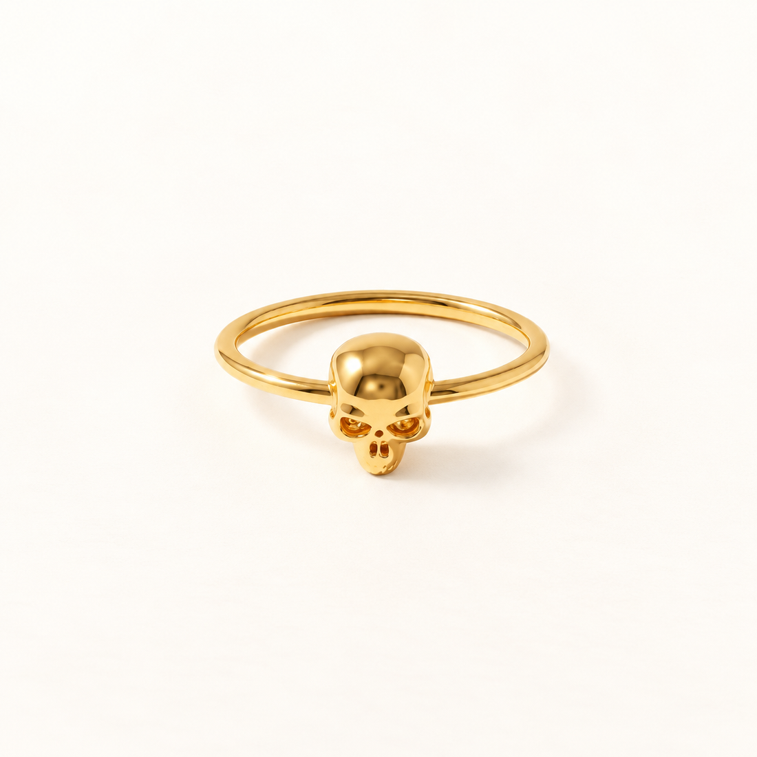 A gold skull ring with delicate band and small detailed skull centerpiece featuring hollow eye sockets and defined jawline, shown from front and side angles on a clean white background displaying the minimalist band, edgy yet feminine aesthetic, and polished gold finish.