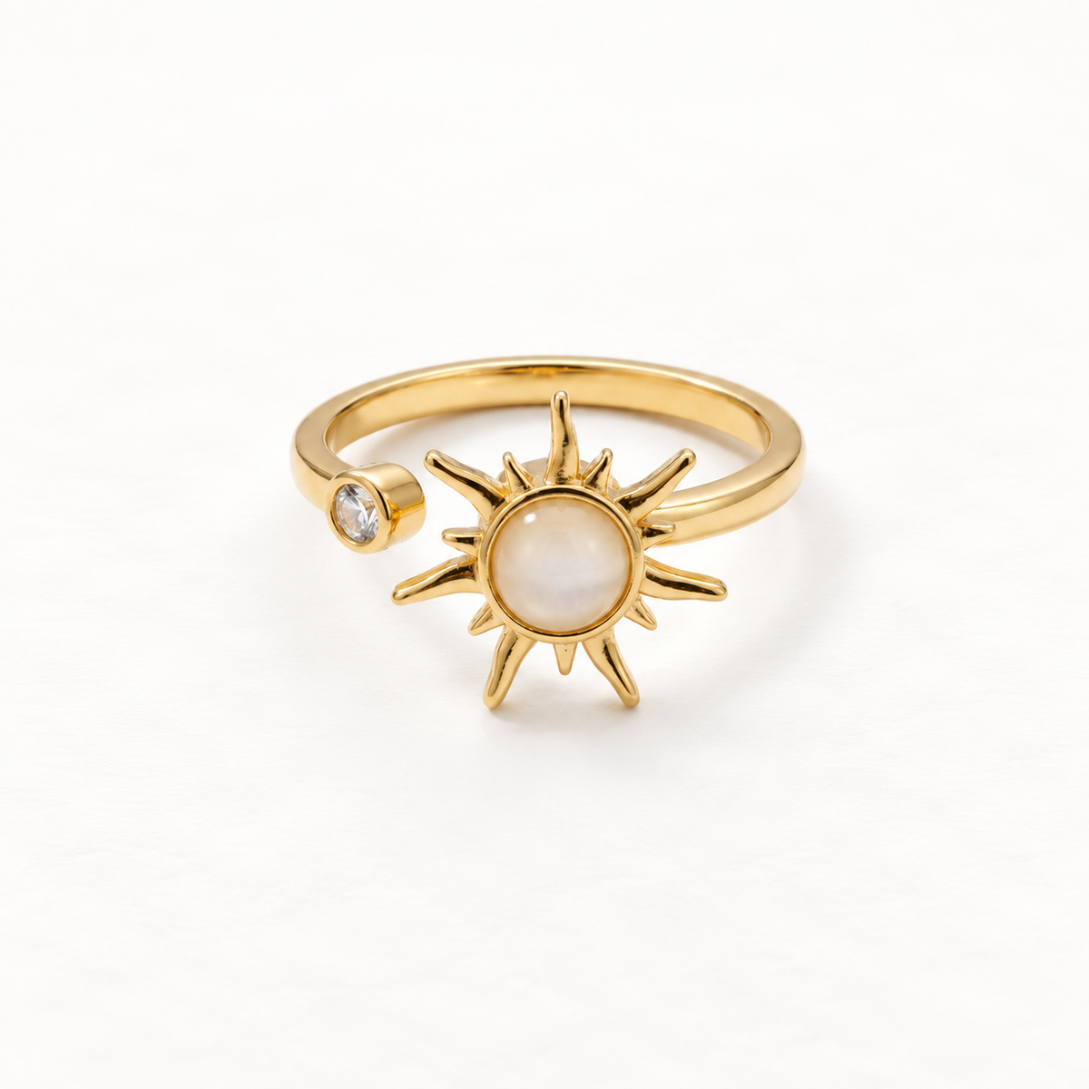 A gold sun ring with iridescent mother of pearl center stone and radiating rays, small clear crystal accent on band, open adjustable design, shown from front and side angles on a clean white background displaying the luminous pearl center, celestial symbolism, and polished gold finish.