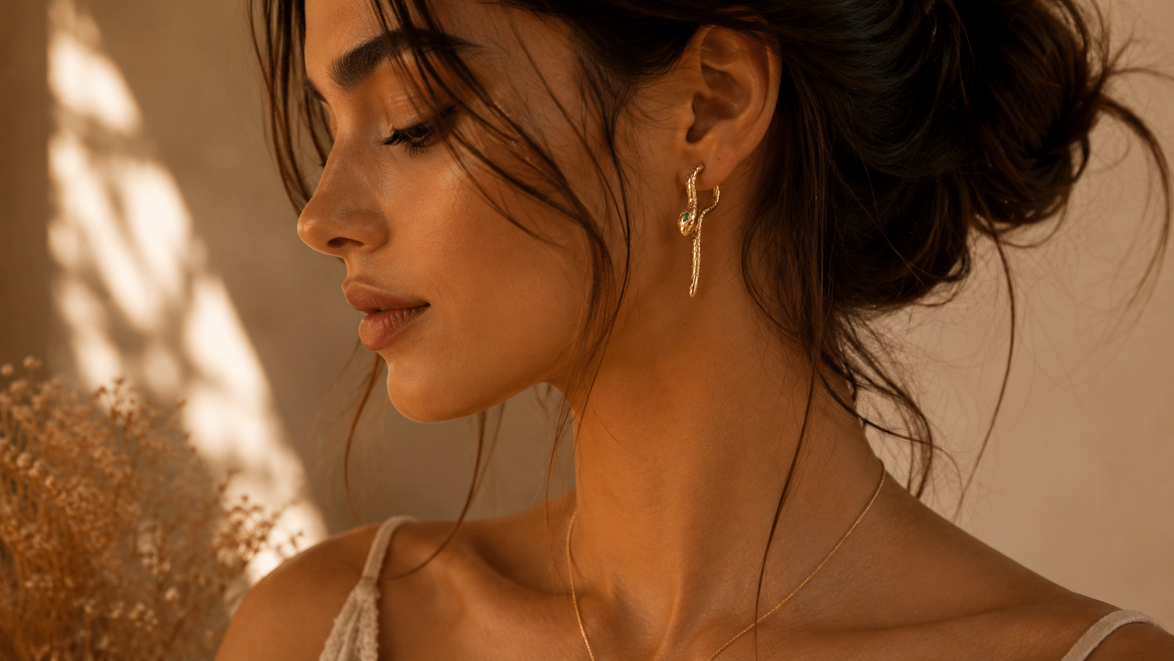 Side profile portrait of woman wearing gold snake earrings with emerald eyes in warm minimalist lighting and soft feminine styling.