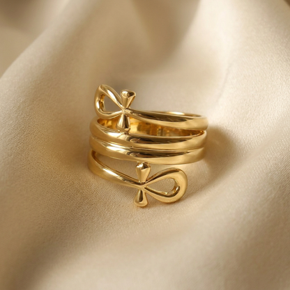 A gold ankh cross ring with Egyptian symbol design featuring looped top and horizontal bar lying flat on a white background with soft natural shadows, showing the ancient hieroglyphic symbol, open adjustable band, and polished gold finish from above.