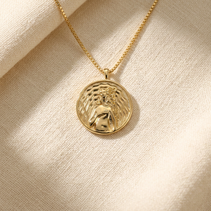 A gold Aphrodite love coin necklace with detailed medallion pendant and box chain lying flat on a white background with soft natural shadows, showing the intricate love symbolism, classical figural relief, and polished gold finish from above.