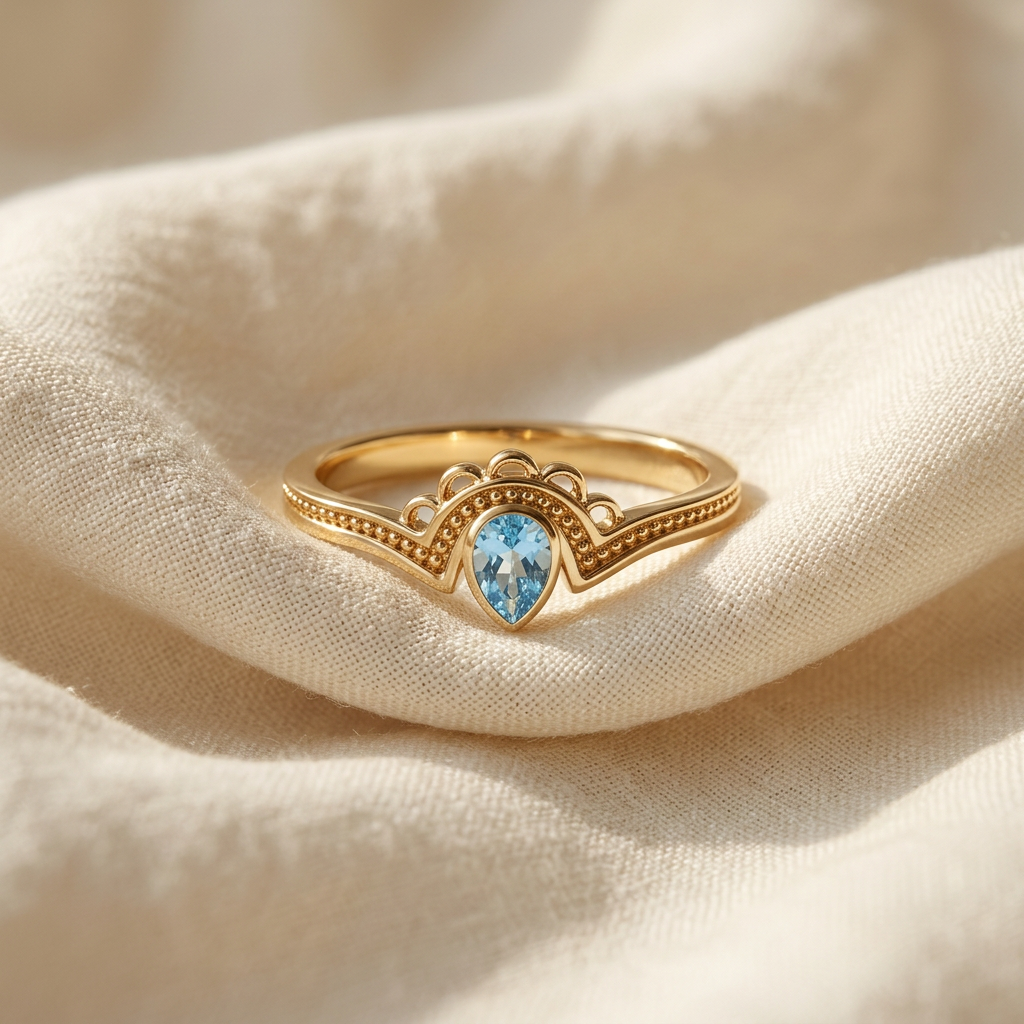 A gold aquamarine teardrop ring with pear-shaped blue stone in vintage-inspired milgrain setting lying flat on a white background with soft natural shadows, showing the soft blue hue, intricate beaded band detail, and polished gold finish from above.