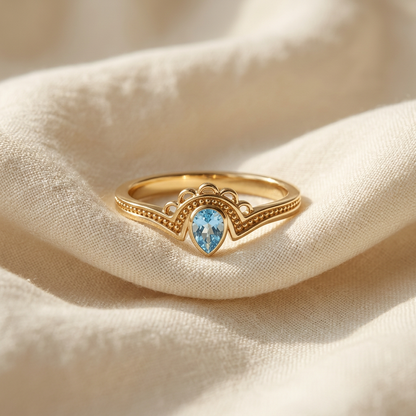 A gold aquamarine teardrop ring with pear-shaped blue stone in vintage-inspired milgrain setting lying flat on a white background with soft natural shadows, showing the soft blue hue, intricate beaded band detail, and polished gold finish from above.