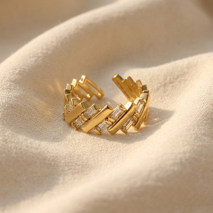 A gold baguette crystal ring with rectangular clear stones set in angular geometric pattern lying flat on a white background with soft natural shadows, showing the sparkling step-cut stones, sleek linear arrangement, and polished gold finish from above.