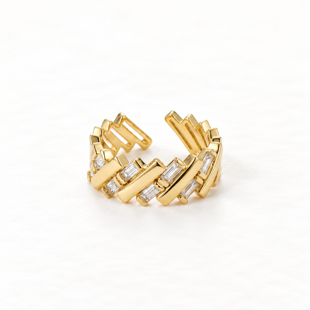 A gold baguette crystal ring with rectangular clear stones set in angular geometric pattern, modern architectural open band design, shown from front and side angles on a clean white background displaying the sparkling step-cut stones, sleek linear arrangement, and polished gold finish.