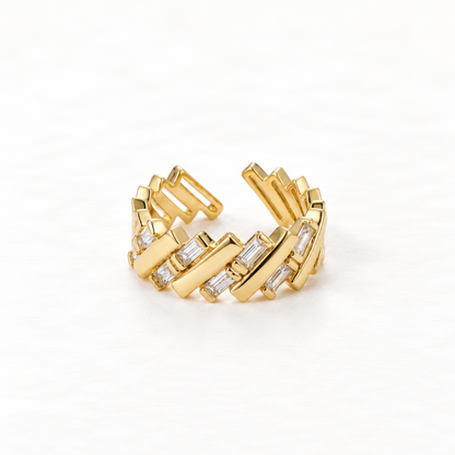 A gold baguette crystal ring with rectangular clear stones set in angular geometric pattern, modern architectural open band design, shown from front and side angles on a clean white background displaying the sparkling step-cut stones, sleek linear arrangement, and polished gold finish.