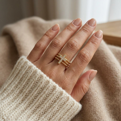 Close-up of a feminine hand wearing a gold beaded cross pave ring with three rows of ball chain bands and crystal-encrusted cross, the delicate sparkling design resting on the finger. Soft natural lighting, warm beige linen background, natural skin texture with visible pores and slight imperfections.
