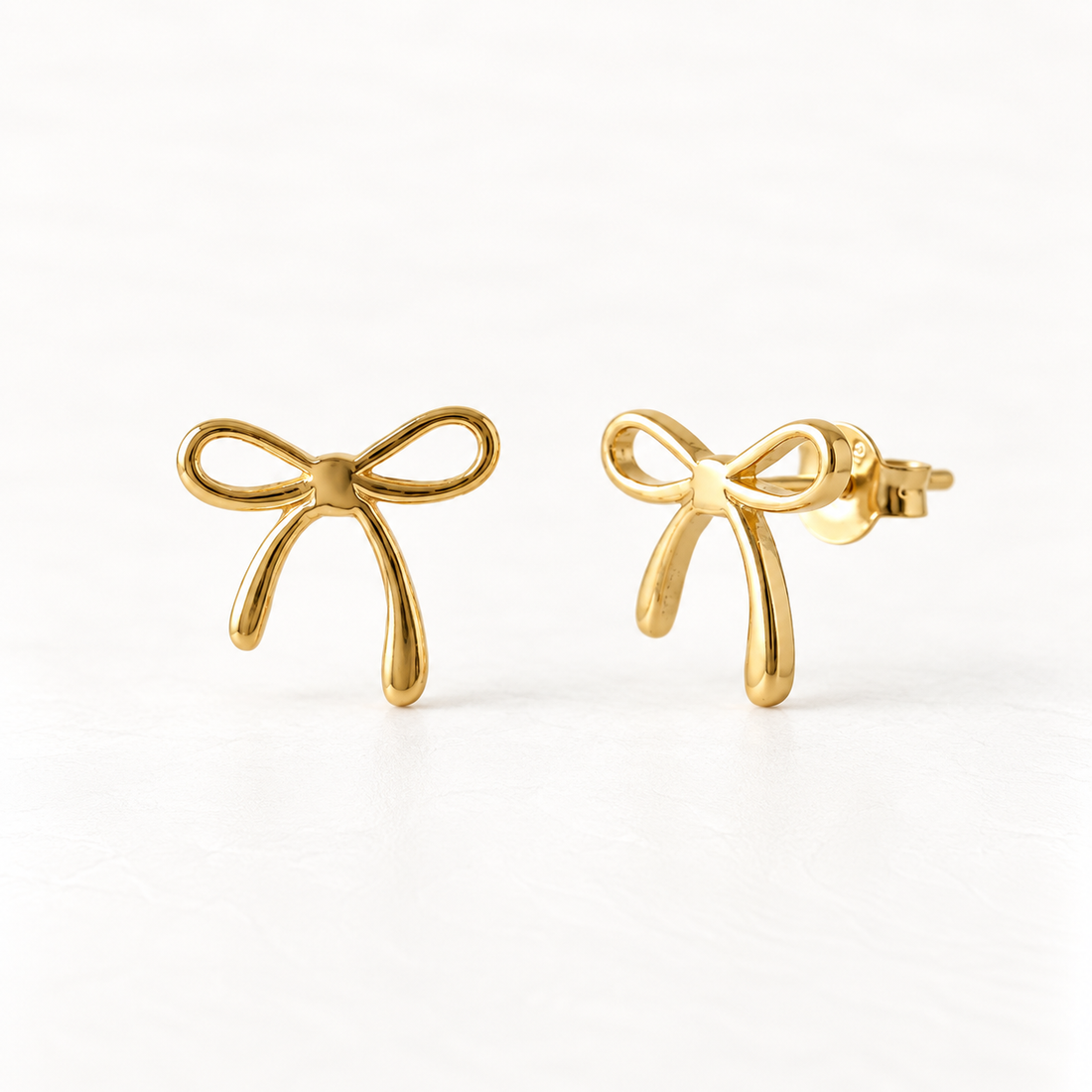 A pair of 18k gold bow stud earrings on a clean white background, one facing forward showing the delicate ribbon bow detail, the other angled to display the post and butterfly backing.