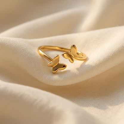A gold butterfly ring with two delicate butterfly silhouettes on open adjustable band lying flat on a white background with soft natural shadows, showing the nature-inspired design, intricate wing texture, and polished gold finish from above.