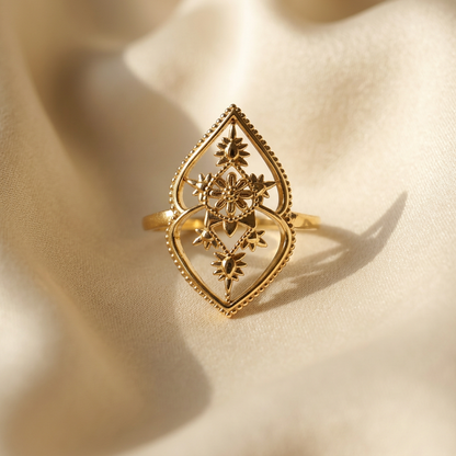 A gold celestial mandala ring with intricate filigree geometric pattern inside diamond-shaped frame lying flat on a white background with soft natural shadows, showing the starburst center, delicate metalwork, and polished gold finish from above.
