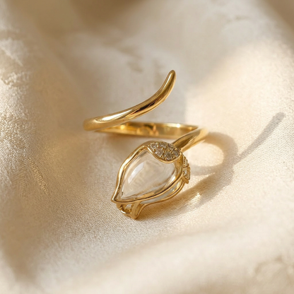 A gold clear crystal teardrop ring with marquise-shaped clear quartz stone in delicate prong setting lying flat on a white background with soft natural shadows, showing the transparent clarity, elegant minimal setting, and polished gold finish from above.