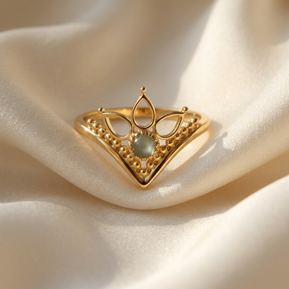 A gold crown tiara ring with intricate filigree details and iridescent moonstone center lying flat on a white background with soft natural shadows, showing the royal symbolism, ornate lace-like texture, and polished gold finish from above.