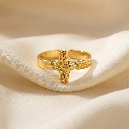 A gold crucifix cross ring with detailed textured 3D cross design and organic hammered band lying flat on a white background with soft natural shadows, showing the sculptural crucifix form, intricate surface texture, and polished gold finish from above.