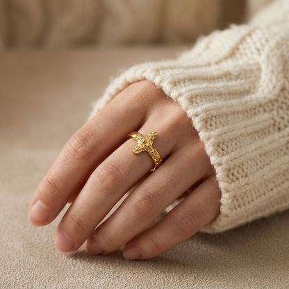 Close-up of a feminine hand wearing a gold crucifix cross ring with detailed textured 3D cross design, the sculptural religious statement resting on the finger. Soft natural lighting, warm beige linen background, natural skin texture with visible pores and slight imperfections.