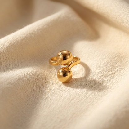 A gold double ball ring with two large polished spherical orbs on open adjustable band lying flat on a white background with soft natural shadows, showing the sculptural spheres, sleek reflective surface, and polished gold finish from above.
