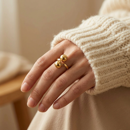 Close-up of a feminine hand wearing a gold double ball ring with two large polished spherical orbs, the modern minimalist statement resting on the finger. Soft natural lighting, warm beige linen background, natural skin texture with visible pores and slight imperfections.