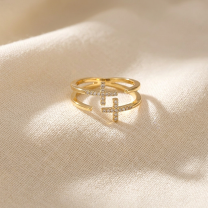 A gold double cross pave ring with two intersecting crosses covered in clear crystals lying flat on a white background with soft natural shadows, showing the sparkling stone detail, geometric symbolism, and polished gold finish from above.