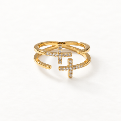 A gold double cross pave ring with two intersecting crosses covered in clear crystals, plus and minus symbol design, open adjustable band, shown from front and side angles on a clean white background displaying the sparkling stone detail, geometric symbolism, and polished gold finish.
