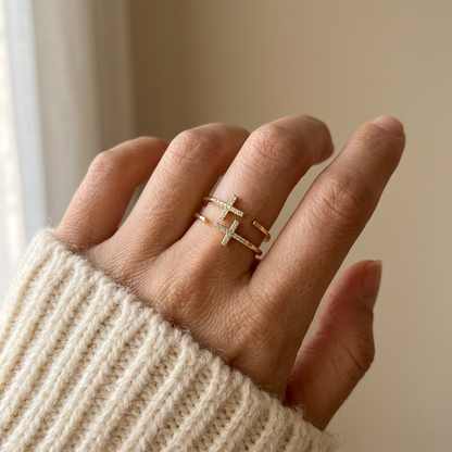 Close-up of a feminine hand wearing a gold double cross pave ring with two intersecting crosses covered in clear crystals, the geometric plus and minus design resting on the finger. Soft natural lighting, warm beige linen background, natural skin texture with visible pores and slight imperfections.