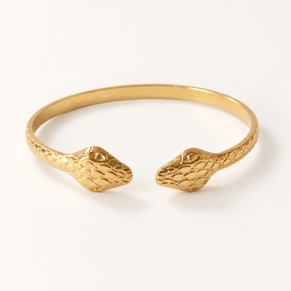 A gold double-headed snake bangle cuff with textured scale pattern and detailed serpent heads on both ends, shown from front and side angles on a clean white background displaying the open cuff design and intricate scale texture.