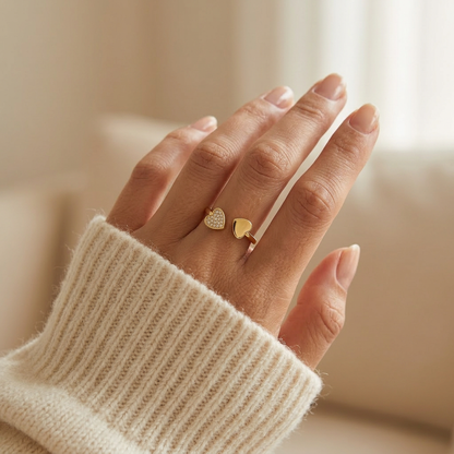 Close-up of a feminine hand wearing a gold double heart ring with one pave crystal heart and one plain heart, the asymmetrical romantic design resting on the finger. Soft natural lighting, warm beige linen background, natural skin texture with visible pores and slight imperfections.