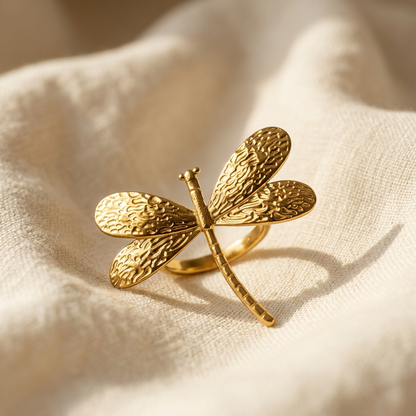 A gold dragonfly ring with detailed veined wing texture and segmented body lying flat on a white background with soft natural shadows, showing the intricate wing pattern, organic body texture, and polished gold finish from above.