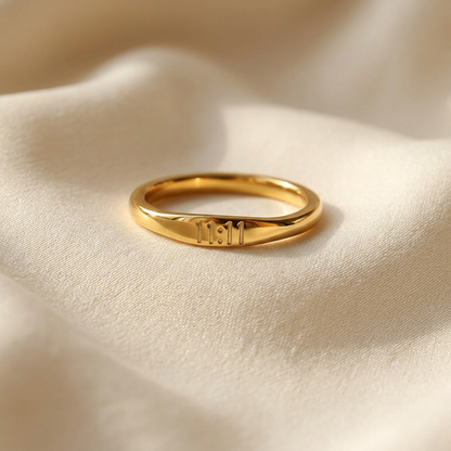 A gold angel number ring with "11:11" engraved on the band lying flat on a white background with soft natural shadows, showing the etched numerals, slim minimalist design, and polished gold finish from above.