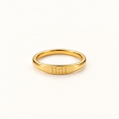 A gold angel number ring with "11:11" engraved on the band, slim minimalist design with etched numerals, shown from front and side angles on a clean white background displaying the spiritual synchronicity symbol, delicate band, and polished gold finish.