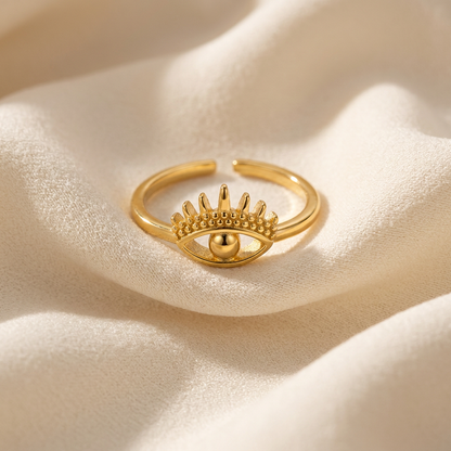 A gold evil eye ring with detailed eyelashes, curved brow, and central spherical pupil lying flat on a white background with soft natural shadows, showing the protective symbol design, intricate lash detail, and polished gold finish from above.