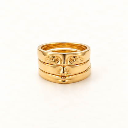 A gold face ring with surrealist facial features including eyes, nose, and mouth across stacked bands, open adjustable design, shown from front and side angles on a clean white background displaying the artistic anthropomorphic detail, sculptural expression, and polished gold finish.