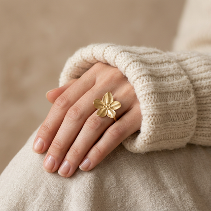 Close-up of a feminine hand wearing a gold five petal flower ring with detailed vein texture, the botanical bloom resting on the finger. Soft natural lighting, warm beige linen background, natural skin texture with visible pores and slight imperfections.