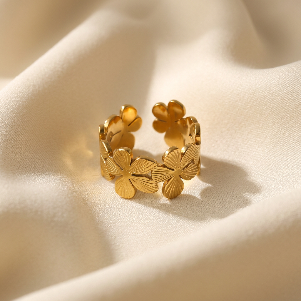 A gold flower band ring with multiple four-petal blossoms encircling the open adjustable band lying flat on a white background with soft natural shadows, showing the continuous floral garden design, intricate botanical texture, and polished gold finish from above.