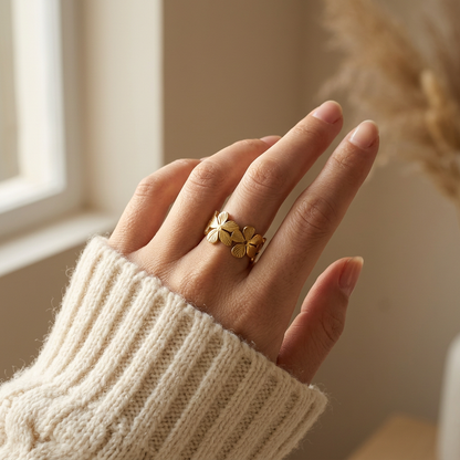 Close-up of a feminine hand wearing a gold flower band ring with multiple four-petal blossoms, the continuous floral garden design resting on the finger. Soft natural lighting, warm beige linen background, natural skin texture with visible pores and slight imperfections.