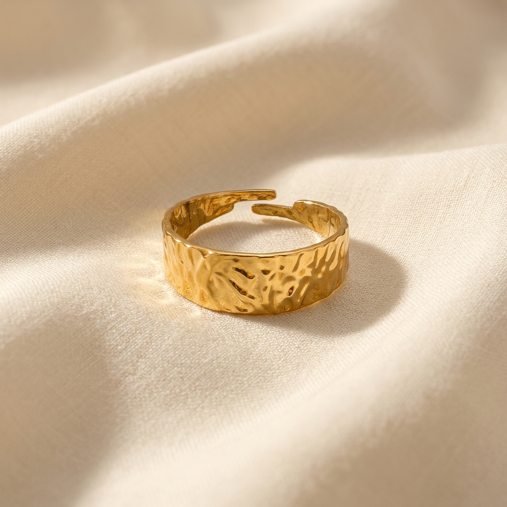 A gold hammered band ring with wide textured surface and organic dimpled pattern lying flat on a white background with soft natural shadows, showing the artisan hammered texture, substantial presence, and polished gold finish from above.