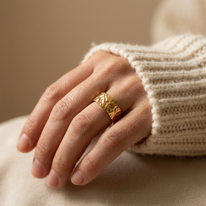 Close-up of a feminine hand wearing a gold hammered band ring with wide textured surface and organic dimpled pattern, the artisan design resting on the finger. Soft natural lighting, warm beige linen background, natural skin texture with visible pores and slight imperfections.