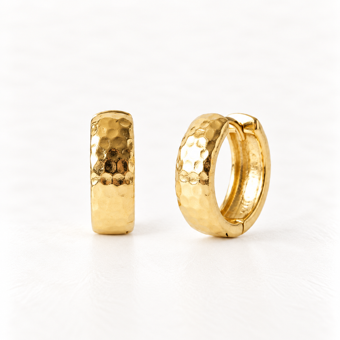 A pair of 18k gold hammered huggie hoop earrings with textured dimpled surface, shown from front and side angles on a clean white background displaying the artisan hammered detail and hinged closure.