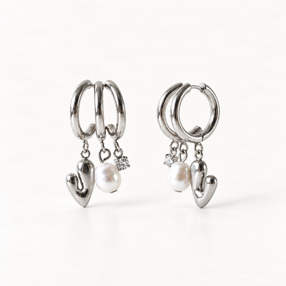A pair of sterling silver huggie hoop earrings with dangling polished heart charms and white freshwater pearl accents, shown from front and side angles on a clean white background displaying the romantic charm detail and hinged closure.
