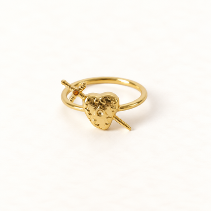 A gold heart and sword ring with detailed anatomical heart pierced by a dagger, open adjustable band, shown from front and side angles on a clean white background displaying the romantic yet edgy symbolism, intricate heart texture and blade detail, and polished gold finish.