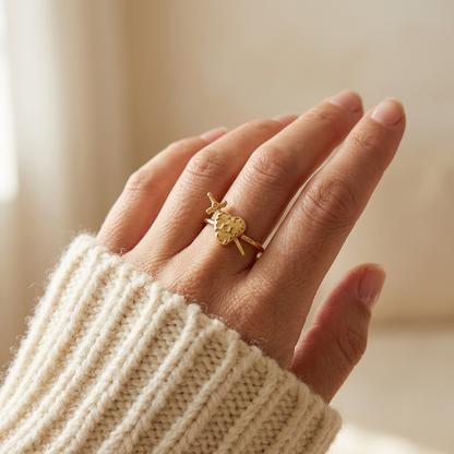 Close-up of a feminine hand wearing a gold heart and sword ring with detailed anatomical heart pierced by a dagger, the romantic yet edgy symbolism resting on the finger. Soft natural lighting, warm beige linen background, natural skin texture with visible pores and slight imperfections.